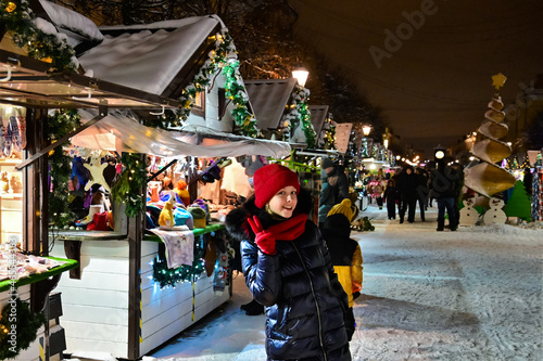 pretty young girl in red hat, scarf and gloves shows victory sign "V" with her fingers against background of winter New Year's fair. Buying gifts for Christmas