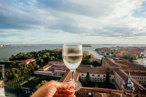 Glass of white wine in hand of a person watching the San Giorgio Maggiore island, Italy. Panorama of Venice lagoon, and gardens under blue sky