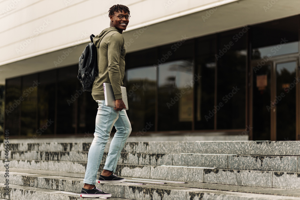 Happy smiling african american student with backpack and laptop on university background