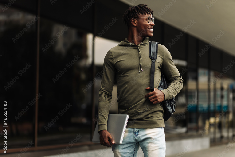Handsome smiling dark-skinned student, wearing glasses with a backpack, holding a laptop, walking around the campus