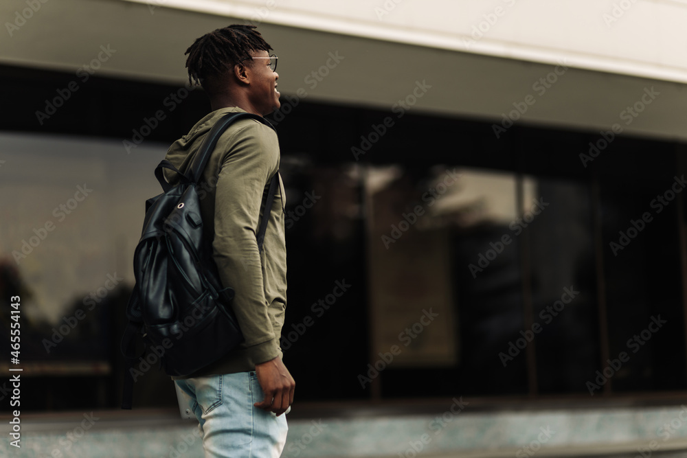 Happy successful african student, wearing glasses and with a black backpack, standing on the steps and smiling