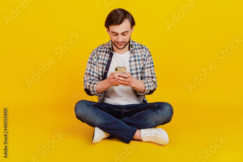 Full length photo of positive cheerful man sit floor use cell phone read news typing sms isolated yellow color background © Tetiana