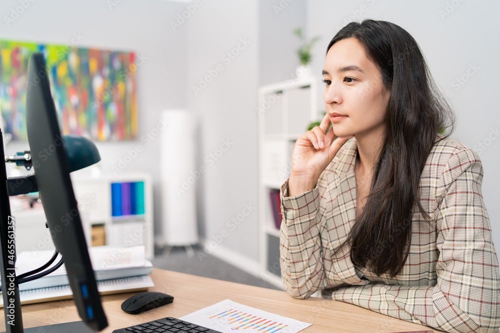 A focused pretty woman with long brown hair wearing a checkered jacket sits at desk in office in front of computer screen, reading news, articles, documents, analyzing propping hand under chin A focused pretty woman with long brown hair wearing a checkered jacket sits at desk in office in front of computer screen, reading news, articles, documents, analyzing propping hand under chin