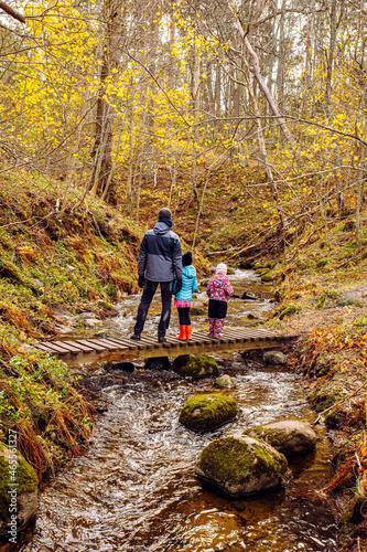 Father hiking with two daughters in beautiful nature in cold autumn day, standing on simple wood bridge. Rough riverbed with hills on background.