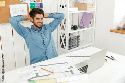 Young white man smiling while working with laptop and drawings in office