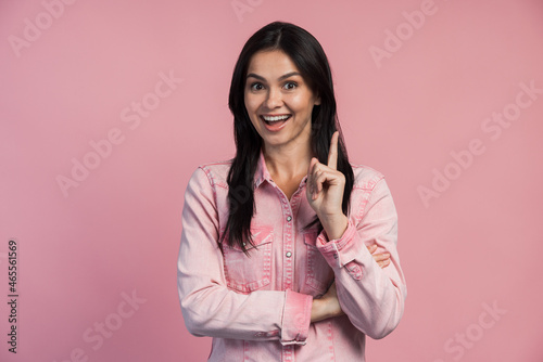 Overjoyed happy woman pointing finger up and looking at camera with toothy smile. Having idea, business plan, startup concept. Indoor studio shot isolated on pink background