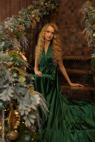 a beautiful blonde with long wavy hair in a chic emerald long evening dress is sitting on the steps of a staircase decorated with Christmas toys