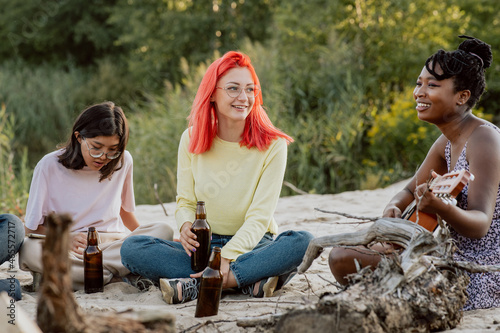 Beautiful black woman with dreadlocks tied up in bun is sitting on sand by lake by fire playing guitar next to friends are listening to the talented woman, drinking beer, spending time together