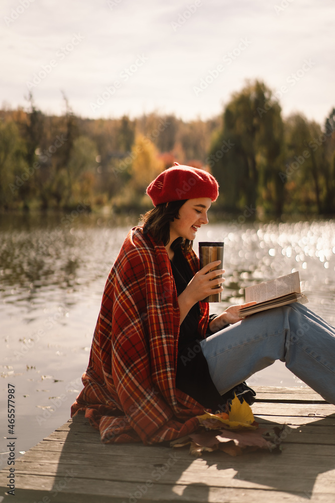 A Teengirl in a red beret reading book on wooden pontoon. Autumn season. Female with trendy clothes relaxing near water reading book and drink coffee. Autumn mood. Autumn concept. Autumn day.
