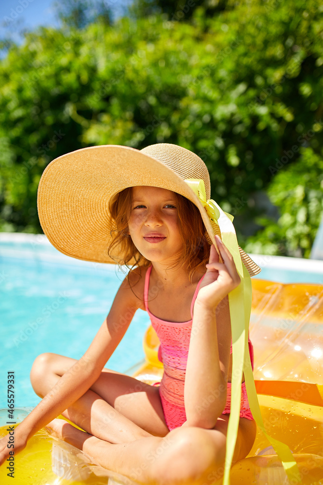 Portrait of little girl in hat relaxing in swimming pool, swims on inflatable yellow mattress