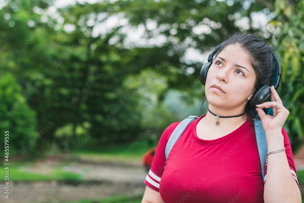 beautiful blue-haired college girl in a red dress listening to music with her wireless headphones in a park, looking up at the sky thoughtfully as she changes songs with her hand on her headphones. beautiful blue-haired college girl in a red dress listening to music with her wireless headphones in a park, looking up at the sky thoughtfully as she changes songs with her hand on her headphones.