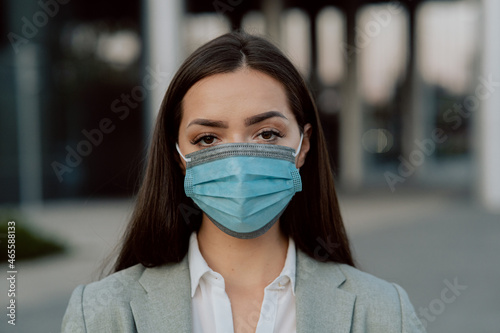 A brunette with dark eyes stands against the background of a city, a glass building, a woman dressed in elegant clothes, a protective mask on face, work, life during the virus pandemic