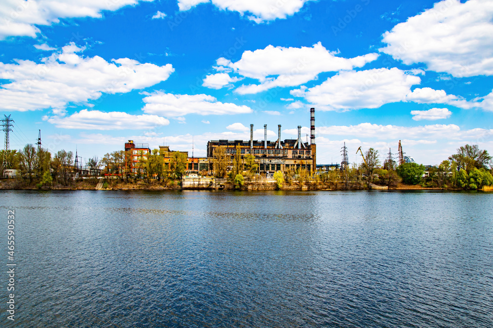 Industrial plant with chimneys on the river bank. Industrial building. Factory workshop. River water flow. Blue sky with clouds. Production approaches. Environmental pollution. Ecology. Business.