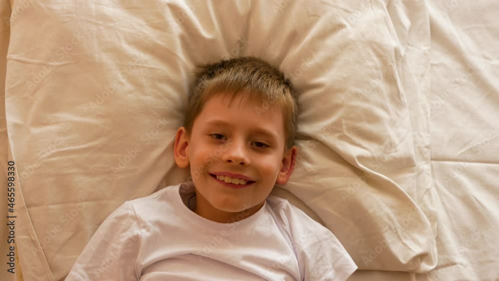 The child wakes up in the morning theme. Positive Caucasian boy 7-9 lying on the bed stretching his arms after sleep and smiling. A carefree person starts a new day. Schoolboy going to school The child wakes up in the morning theme. Positive Caucasian boy 7-9 lying on the bed stretching his arms after sleep and smiling. A carefree person starts a new day. Schoolboy going to school