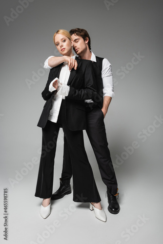 full length view of elegant man embracing blonde woman while standing with hand in pocket on grey