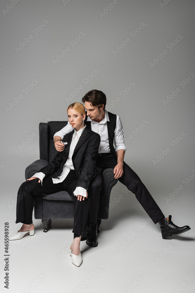 stylish man lightening cigarette of blonde woman while sitting in armchair on grey stylish man lightening cigarette of blonde woman while sitting in armchair on grey