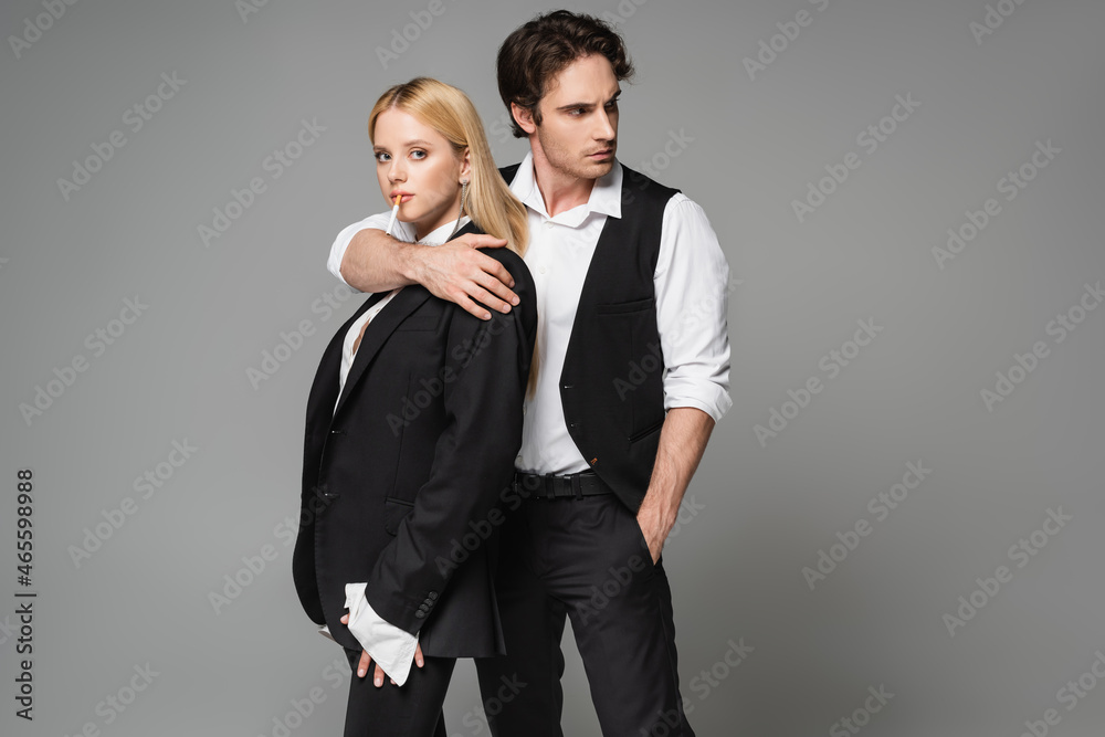 blonde woman with cigarette looking at camera while stylish man embracing her isolated on grey blonde woman with cigarette looking at camera while stylish man embracing her isolated on grey