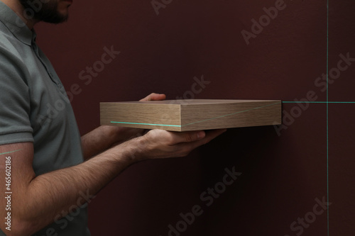 Man using cross line laser level for hanging shelf on brown wall, closeup