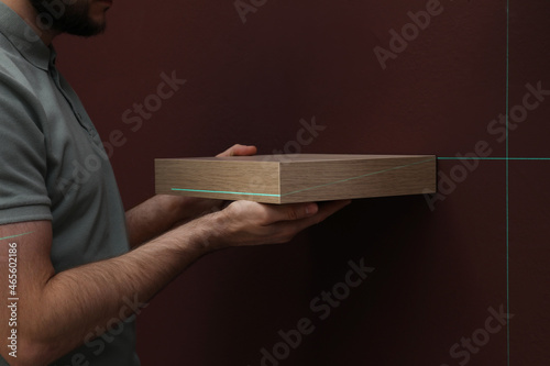 Fotografie Man using cross line laser level for hanging shelf on brown wall, closeup