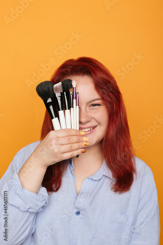 Portrait of young smiling visagist with makeup brushes in hand. Red-haired girl isolated on orange. Professional