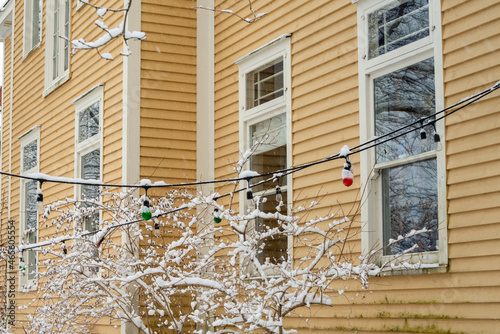 The exterior yellow wall of a wood building with multiple double hung windows. The house and its windows have beige trim. There are multi-colored Christmas lights strung in front of the windows. 