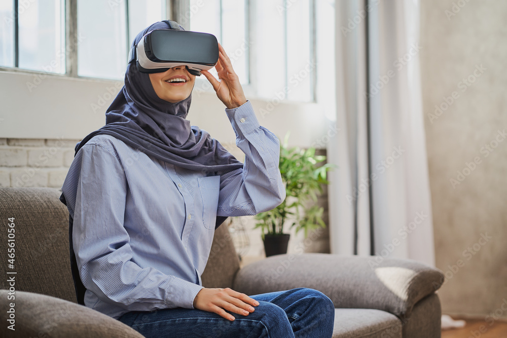 Joyful Arabian woman sitting on a couch, wearing a VR helmet and smiling Joyful Arabian woman sitting on a couch, wearing a VR helmet and smiling