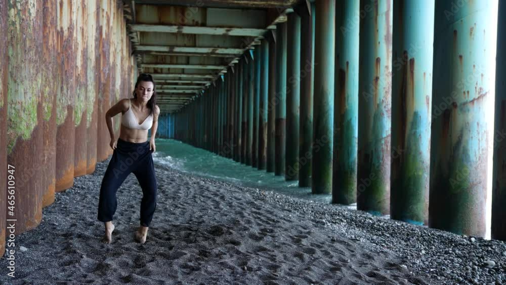 a barefoot woman dances under the pillars of the bridge against the background of the incoming waves. she makes undulating movements with arms and legs. the general plan a barefoot woman dances under the pillars of the bridge against the background of the incoming waves. she makes undulating movements with arms and legs. the general plan