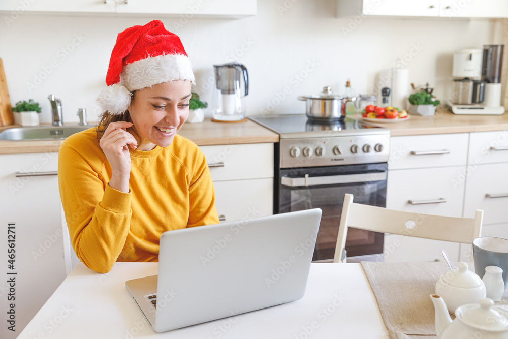 Smiling girl wearing Santa hat having video calling family by webcam. Woman with laptop sitting on kitchen at home using virtual meeting chat on holidays. Happy Christmas and New Year new normal. Smiling girl wearing Santa hat having video calling family by webcam. Woman with laptop sitting on kitchen at home using virtual meeting chat on holidays. Happy Christmas and New Year new normal.