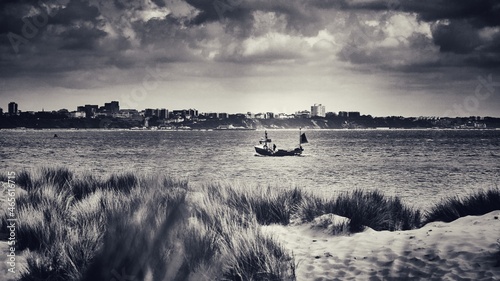 Fishing boat sailing into Poole Harbour with Bournemouth in the background, and Shell Bay sand dunes in the foreground, black and white sepia