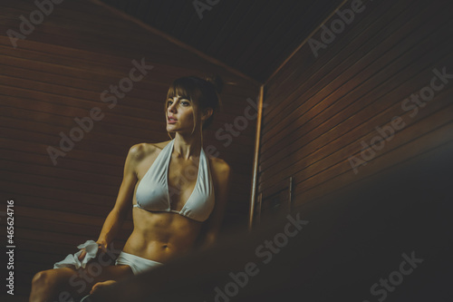Beautiful healthy girl relaxing in a wooden sauna with a white bikini. Young woman resting after workout in the gym. Fitness female with towel, laying on the bench in the spa. Wellness, health concept