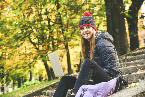 Girl with wool cap sitting in the park using laptop, beautiful autumn afternoon. Teen use computer to communicate with friends and classmates. Young student woman enjoying free time in nature