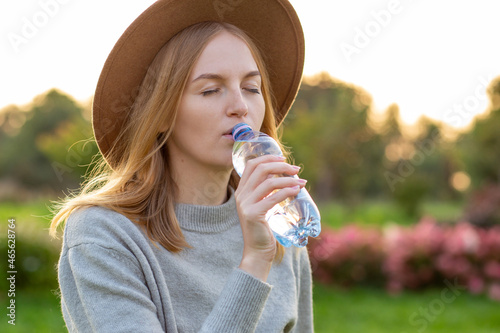 Close up portrait of an attractive young blonde woman in hat drinking water from plastic bottle at summer green park. Food, rest or ecology concept
