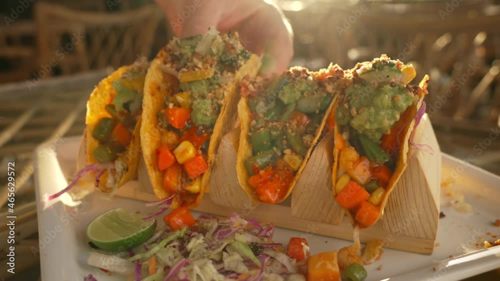 A person takes an appetizing Mexican tacos from a white plate in the rays of warm sunlight. Dinner with tacos with a bright filling and a drink, on a glass table during sunset in a cafe. Close-up.