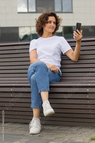 A 45-50-year-old woman is sitting on a bench and taking a selfie on a mobile phone