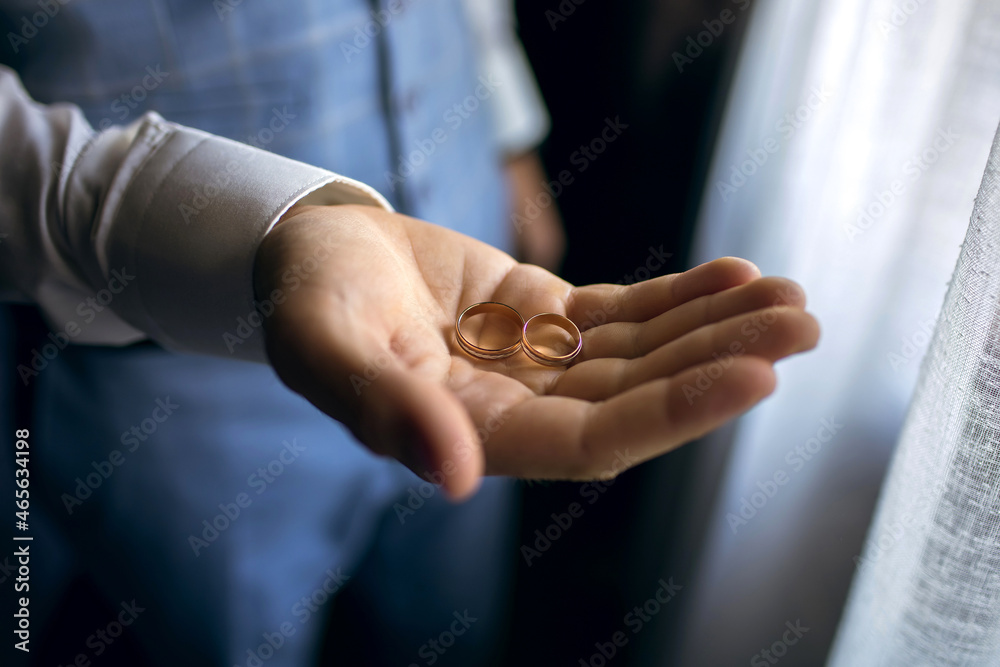 Obraz premium The groom on the wedding day holds two wedding rings in his hand in a hotel room. The man is wearing a white shirt and blue pants with a vest. Gold wedding rings close up on the palm
