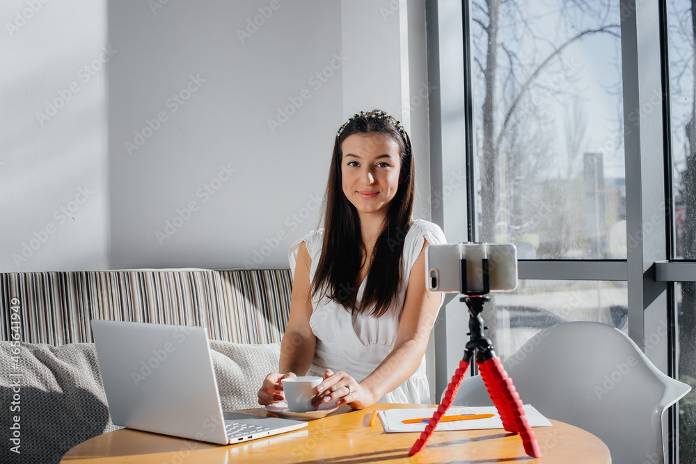 Obraz premium A young beautiful girl is sitting in a cafe, recording video blogs and chatting on social networks.