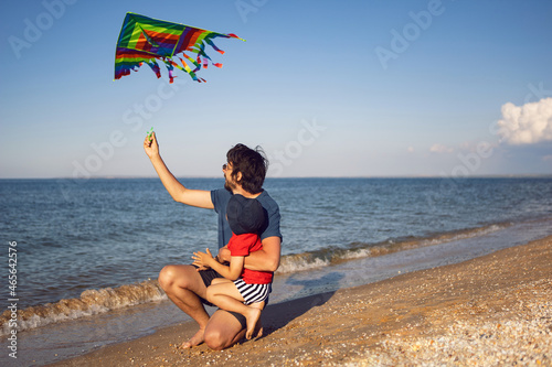 Fototapeta Naklejka Na Ścianę i Meble -  father and son are standing on a sandy beach by the sea and launch toy striped kite in the summer on vacation
