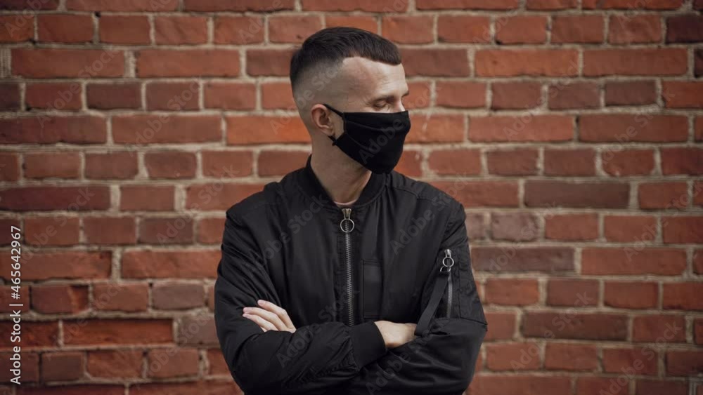 Caucasian serious male security in a black jacket against the background of a brick red wall looks around, takes off and throws away the protective mask, breathes freely putting himself in danger Caucasian serious male security in a black jacket against the background of a brick red wall looks around, takes off and throws away the protective mask, breathes freely putting himself in danger