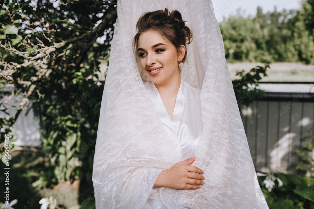 A beautiful, young, sweet brunette bride in a white robe, lingerie stands outdoors in the garden, wrapped in her favorite lace dress hanging on a tree. Portrait, photography.