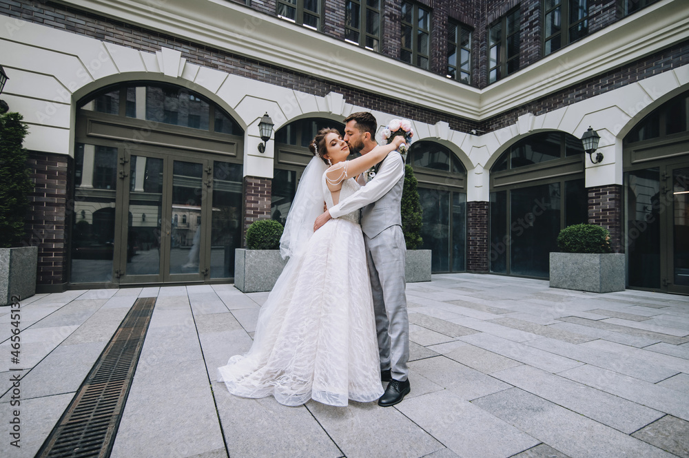 A beautiful bride in a white dress with a long veil and a stylish bearded groom in a gray suit with a bow tie hug and kiss against the backdrop of architecture, red brick houses with windows.