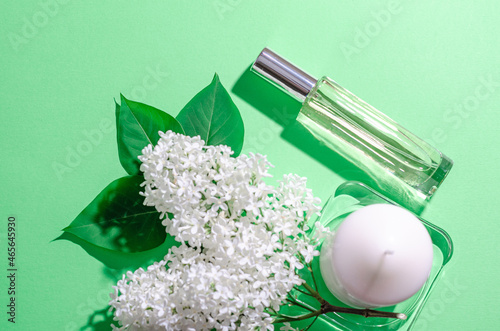 White lilac flowers, white candle and a glass bottle with perfume on a green background. Bright shadows and light. Place for your text. Flat lay. SPA and aromatherapy.