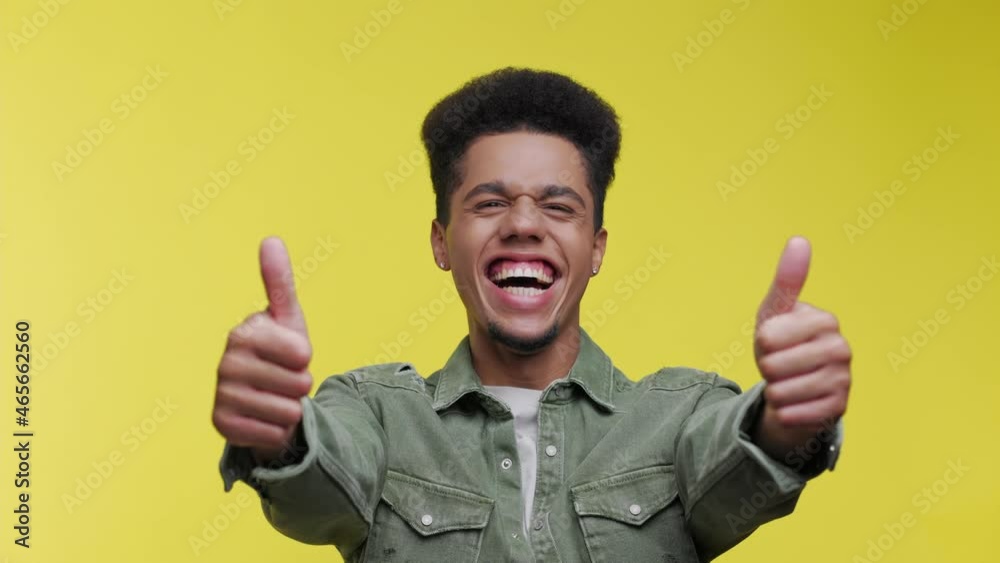 A smiling positive young african american man is doing a thumb-up gesture isolated over yellow wall background in studio. People lifestyle concept. Guy looking camera showing thumbs up. A smiling positive young african american man is doing a thumb-up gesture isolated over yellow wall background in studio. People lifestyle concept. Guy looking camera showing thumbs up.