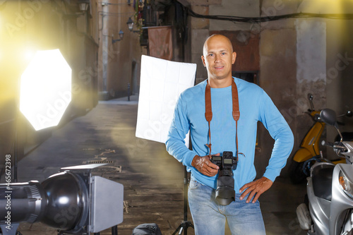 Professional photographer. Portrait of confident adult man holding camera in hands while standing among lighting equipment on city street