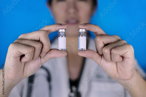 A female doctor holding 2 vials of vaccine in parallel. A physician in white coat and stethoscope around her neck showing 2 glass bottles with blank white labels, comparing them.