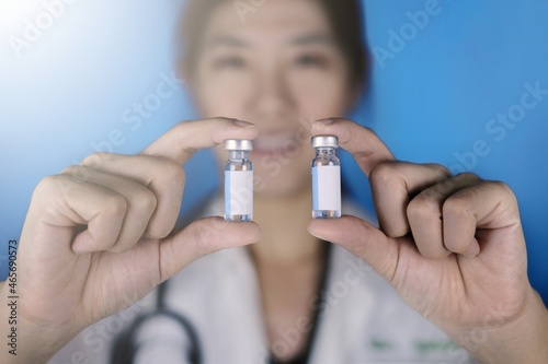 A female doctor holding 2 vials of vaccine in parallel. A physician in white coat and stethoscope around her neck showing 2 glass bottles with blank white labels, comparing them.