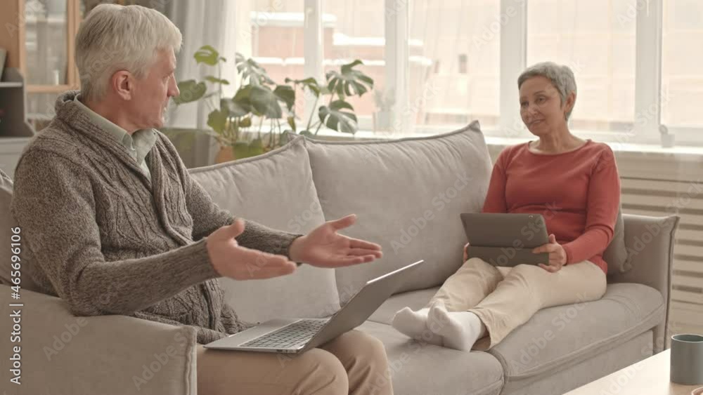 Steadicam of senior Caucasian man typing on laptop on couch, talking to his short-haired Asian wife who sitting with legs up, using tablet computer, smiling Steadicam of senior Caucasian man typing on laptop on couch, talking to his short-haired Asian wife who sitting with legs up, using tablet computer, smiling