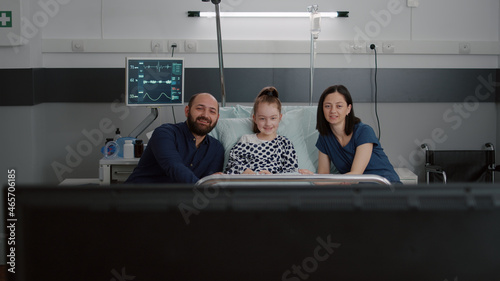 Family watching entertainment cartoon movie on television in hospital ward while waiting for medical expertise during recovery examination. Sick child patient resting in bed after illness surgery