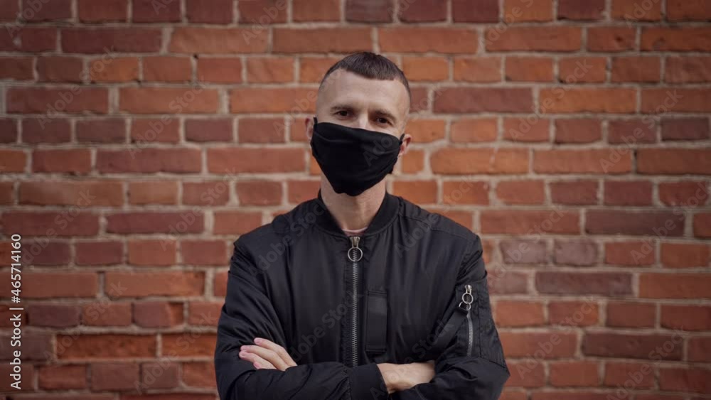 a disgruntled male bodyguard with the appearance of a bully with a short haircut against a red brick wall takes off, throws on camera a protective mask against covid.