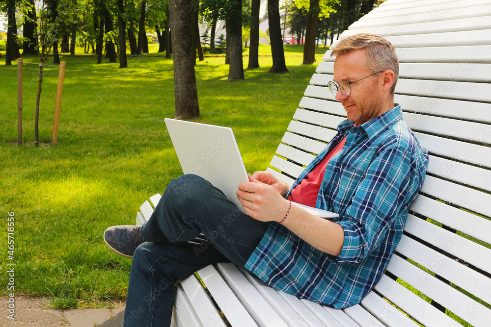 A young man in glasses looks at a laptop in the park. A man sits on a bench with a white laptop. A young man in glasses looks at a laptop in the park. A man sits on a bench with a white laptop.