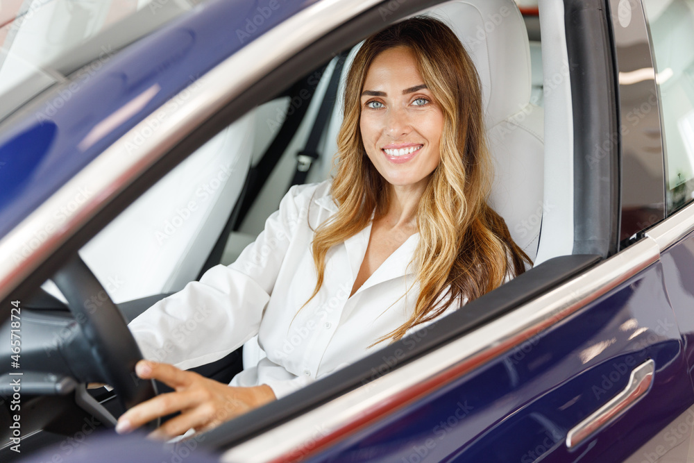 Smiling driver woman customer buyer client in shirt hold put hand on steering wheel choose auto want buy new automobile in car showroom vehicle salon dealership store motor show indoor. Sales concept. Smiling driver woman customer buyer client in shirt hold put hand on steering wheel choose auto want buy new automobile in car showroom vehicle salon dealership store motor show indoor. Sales concept.